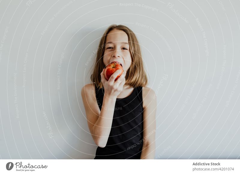 Entzücktes Kind beißt Apfel gegen weiße Wand Mädchen Biss heiter Lächeln essen frisch Frucht Vitamin genießen Porträt Mahlzeit Freude Glück reif lässig Top