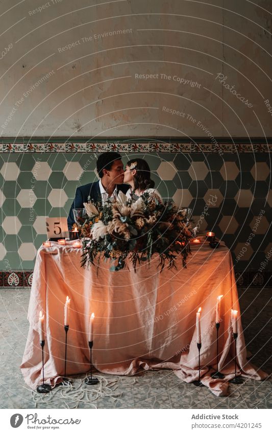 Multiethnisches frisch vermähltes Paar am Tisch mit Dekoration am Hochzeitstag Jungvermählter Kuss Heirat Dekoration & Verzierung Partnerschaft festlich feiern