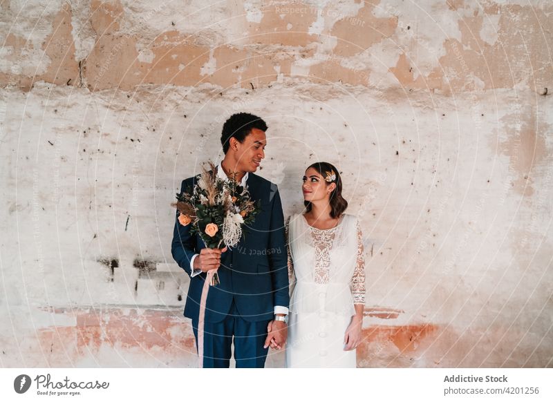 Aufrichtiges multiethnisches, frisch verheiratetes Paar, das sich in der Nähe einer rauen Wand an den Händen hält Händchenhalten Jungvermählter Hochzeit