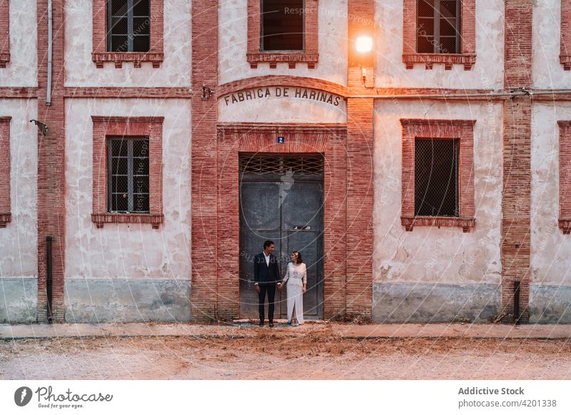 Romantisches ethnisches, frisch verheiratetes Paar, das sich vor einem alten Backsteingebäude die Hände hält Jungvermählter Hochzeit Händchenhalten Angebot