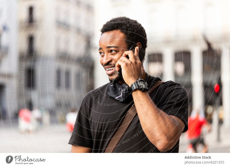 Lächelnder schwarzer Mann, der auf einer Straße in der Stadt mit seinem Smartphone spricht sprechen freundlich maskulin Gespräch Porträt benutzend Apparatur