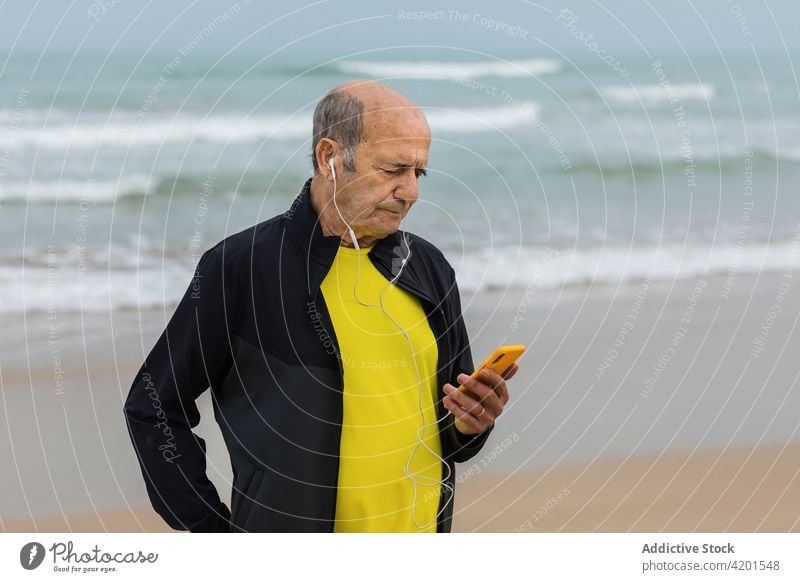 Älterer Sportler beim Musikhören am Meer zuhören MEER Ufer Smartphone benutzend winken Training Pause männlich Fitness älter gealtert Senior Kopfhörer Athlet