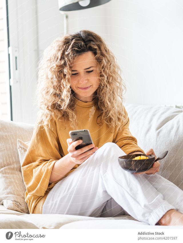Fröhliche Frau, die mit einer Schüssel Essen auf dem Sofa sitzt und auf ihrem Smartphone surft positiv heiter benutzend Textnachricht Appartement Lebensmittel