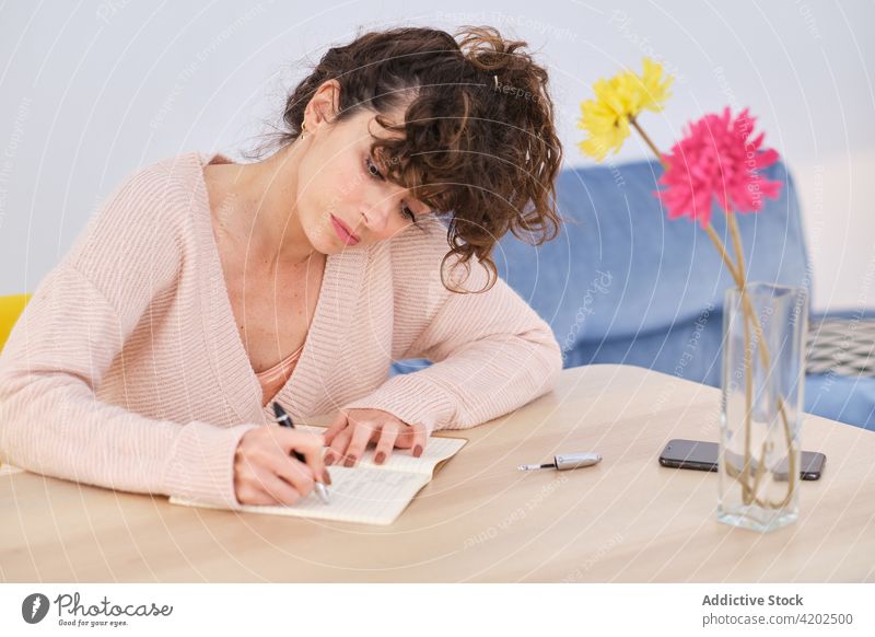 Nachdenkliche Frau schreibt zu Hause Notizen in ein Schreibheft schreiben Notizblock Raum zur Kenntnis nehmen Schönschreibheft Smartphone Vase Blume