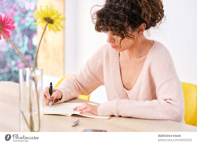 Nachdenkliche Frau schreibt zu Hause Notizen in ein Schreibheft schreiben Notizblock Raum zur Kenntnis nehmen Schönschreibheft Smartphone Vase Blume