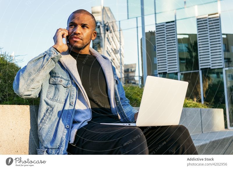 Schwarzer Freiberufler mit Laptop und Smartphone in der Stadt sitzend Mitarbeiter freiberuflich maskulin diskutieren sprechen nachdenken Handy Mann urban