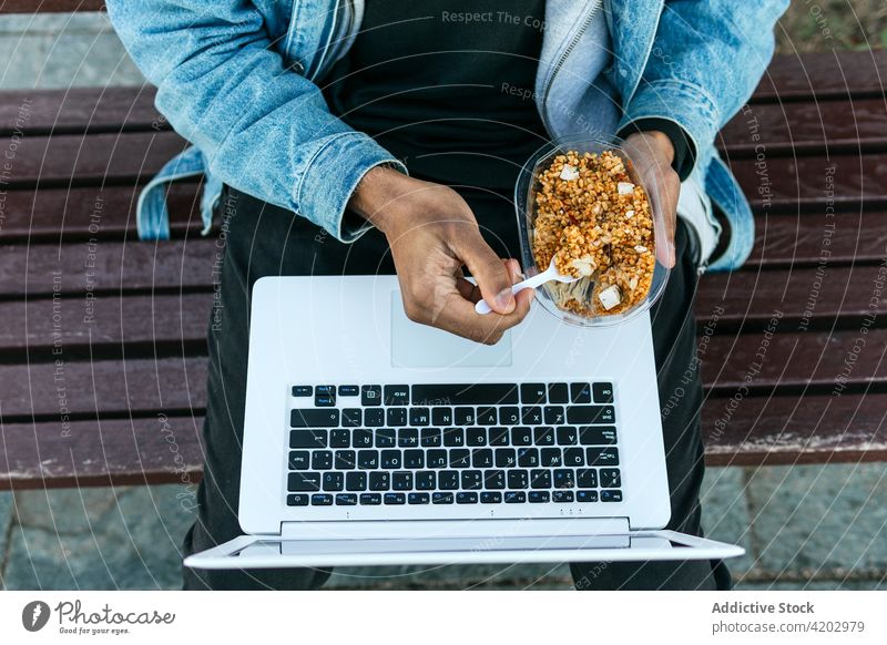 Crop schwarz remote Mitarbeiter mit Laptop essen Essen auf der Straße Lebensmittel Mittagessen lecker verträumt Mann Stadt Arbeiter freiberuflich