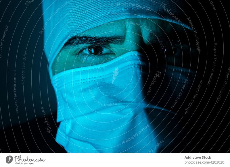 Männlicher Arzt mit Maske in einem dunklen Raum mit blauem Neonlicht Chirurg Mann Mundschutz medizinisch dunkel neonfarbig Licht männlich Medizin behüten