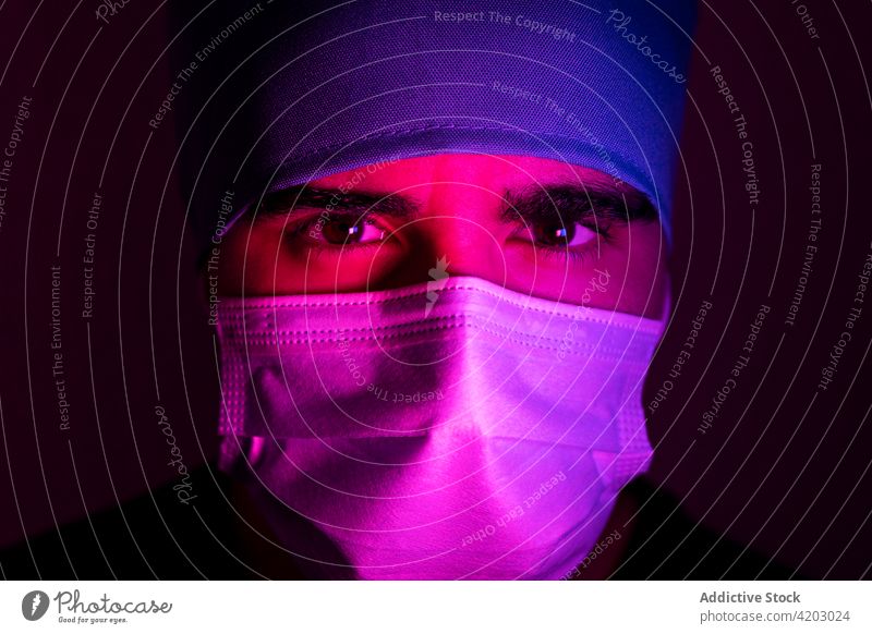 Männlicher Arzt mit Maske in einem dunklen Raum mit blauem und rotem Neonlicht Chirurg Mann Mundschutz medizinisch dunkel neonfarbig Licht Medizin behüten