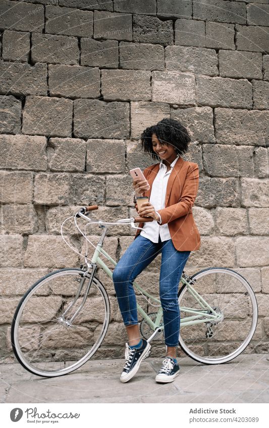 Fröhliche schwarze Frau, die in der Nähe eines Fahrrads in der Stadt auf ihrem Smartphone surft Browsen soziale Netzwerke heiter Großstadt Imbissbude trinken