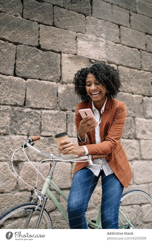 Fröhliche schwarze Frau, die in der Nähe eines Fahrrads in der Stadt auf ihrem Smartphone surft Browsen soziale Netzwerke heiter Großstadt Imbissbude trinken