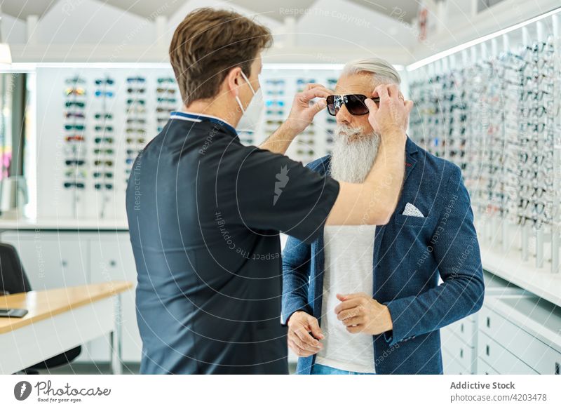 Optiker hilft einem älteren Mann beim Einsetzen einer Sonnenbrille im Optikgeschäft Hilfsbereitschaft optisch Laden Senior angezogen Brille Kunde Dienst