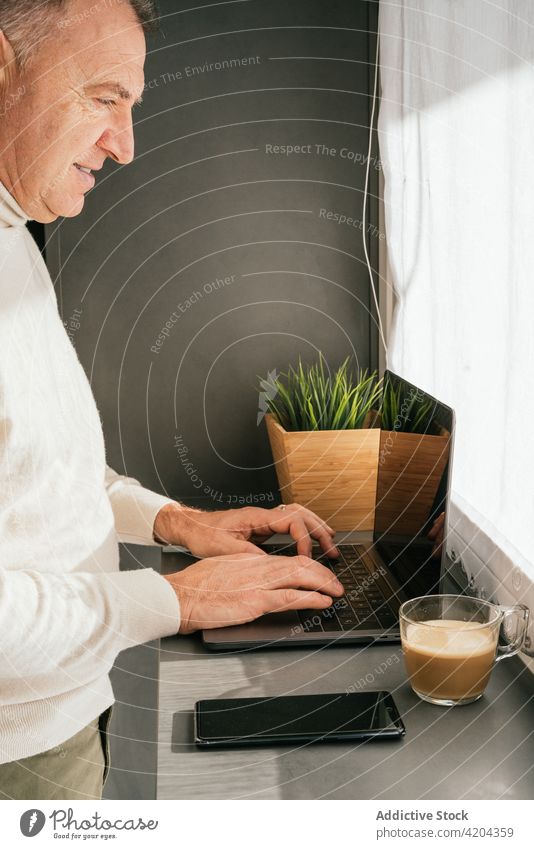 Fröhlicher reifer Mann sitzt auf einem Stuhl am Tresen mit Laptop in der Küche Morgen Lächeln heimwärts Frühstück Kaffee Abfertigungsschalter männlich Arbeit
