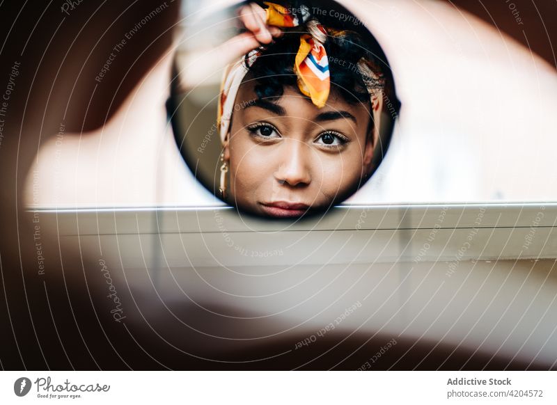 Charmante schwarze Frau mit Stirnband schaut in einen runden Spiegel angezogen charmant Accessoire Kopfbedeckung heimwärts Inhalt ethnisch Afroamerikaner