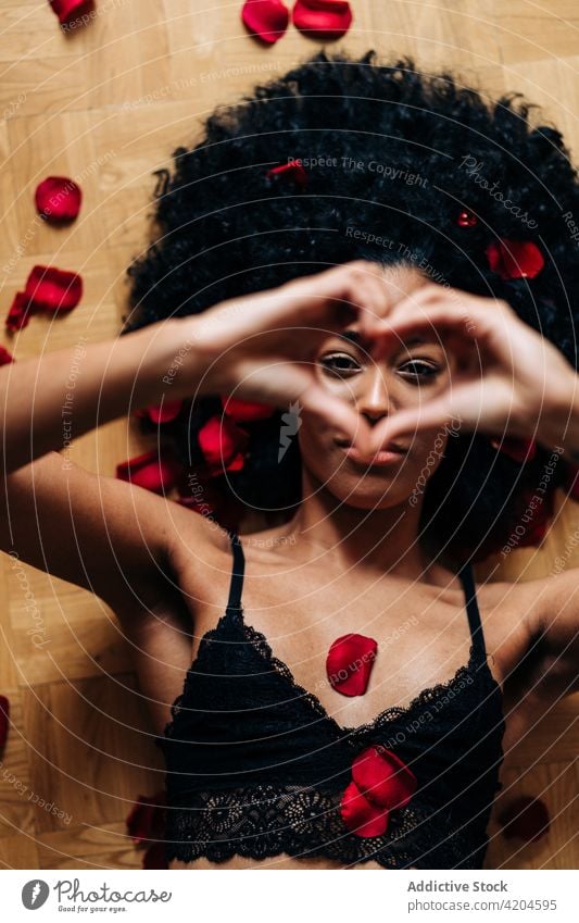 Schwarze Frau zeigt Herzgeste auf dem Boden mit Rosenblättern gestikulieren Liebe Zeichen Roséwein Blütenblatt romantisch Valentinsgruß Angebot ethnisch schwarz