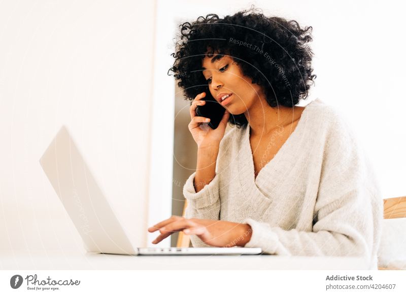 Schwarze Frau, die zu Hause mit ihrem Smartphone spricht und an ihrem Laptop arbeitet multitask freiberuflich heimwärts Arbeit abgelegen Projekt beschäftigt