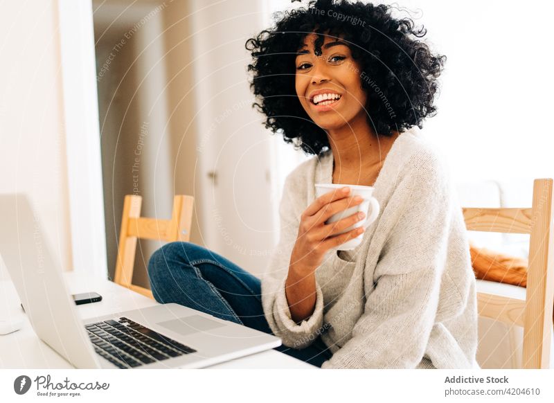 Zufriedene schwarze Frau sitzt am Tisch mit Laptop und Tasse mit Getränk freiberuflich heiter heimwärts Büro trinken Unternehmer Projekt ethnisch Afroamerikaner