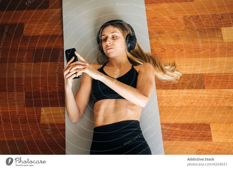 Sportliche Frau mit Kopfhörern, die nach dem Training auf ihrem Mobiltelefon surft Smartphone Browsen zuhören Musik ruhen Unterlage Sportkleidung Übung Athlet