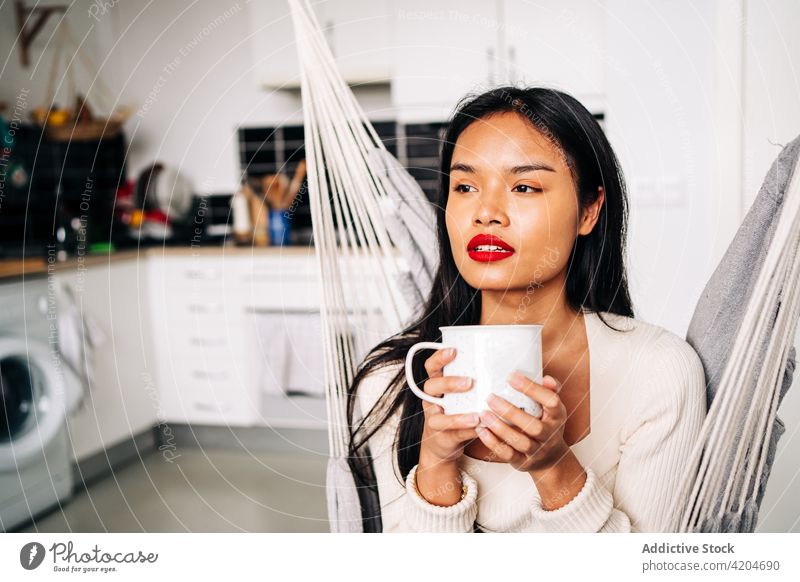 Hispanische Frau sitzt zu Hause in der Hängematte Getränk hispanisch jung heimwärts sitzen trinken Tee Dame Becher Freizeit Tasse Appartement Zeitgenosse
