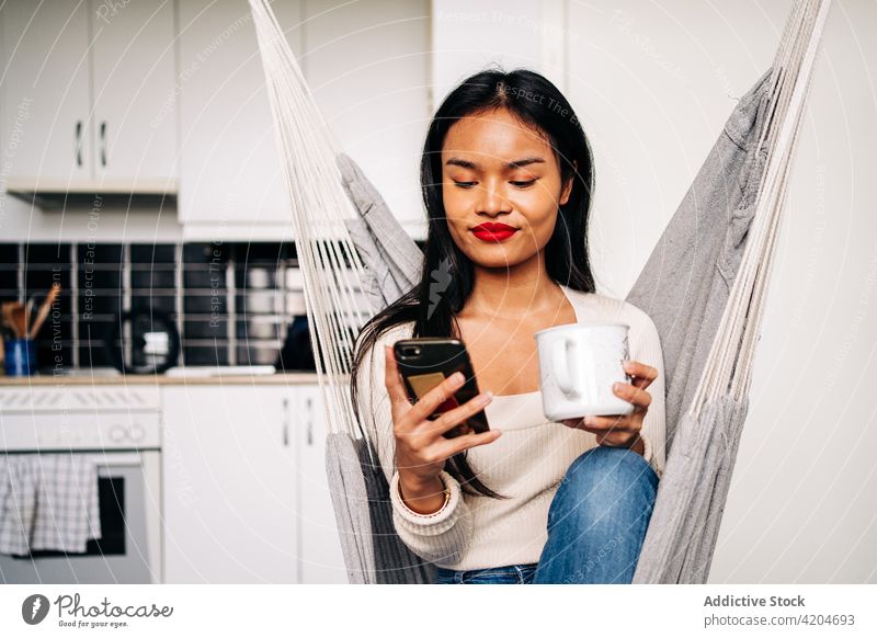 Hispanische Frau, die in der Hängematte sitzend eine SMS über ihr Smartphone verschickt Getränk benutzend soziale Netzwerke online Browsen Gerät hispanisch