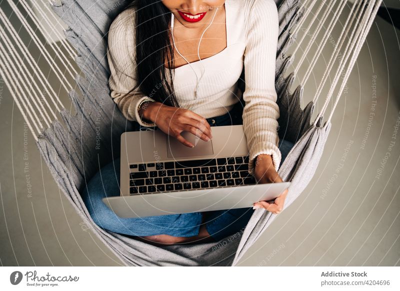 Fröhliche junge Frau, die in der Hängematte sitzend am Laptop arbeitet freiberuflich Lächeln Browsen Apparatur soziale Netzwerke Kopfhörer benutzend Komfort
