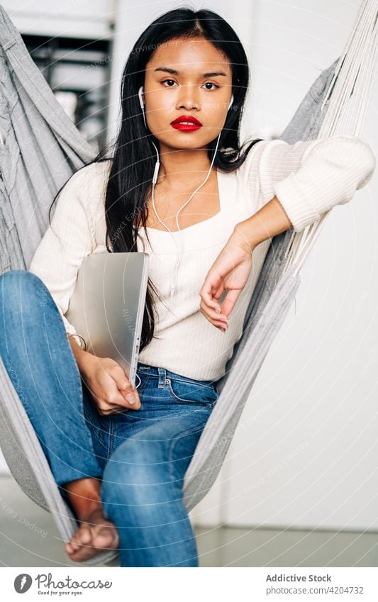Nachdenkliche junge Frau mit Laptop in der Hängematte sitzend freiberuflich Browsen Apparatur Halt soziale Netzwerke nachdenklich Kopfhörer benutzend besinnlich