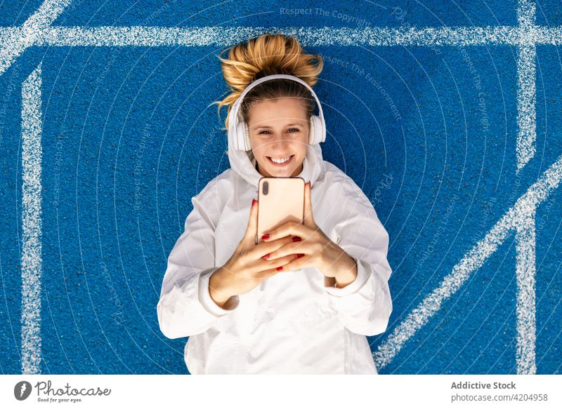 Zufriedene Sportlerin macht Selfie auf der Rennbahn im Stadion Athlet Smartphone Selbstportrait Leichtathletik Kopfhörer Musik Frau Lächeln Lügen blau Farbe