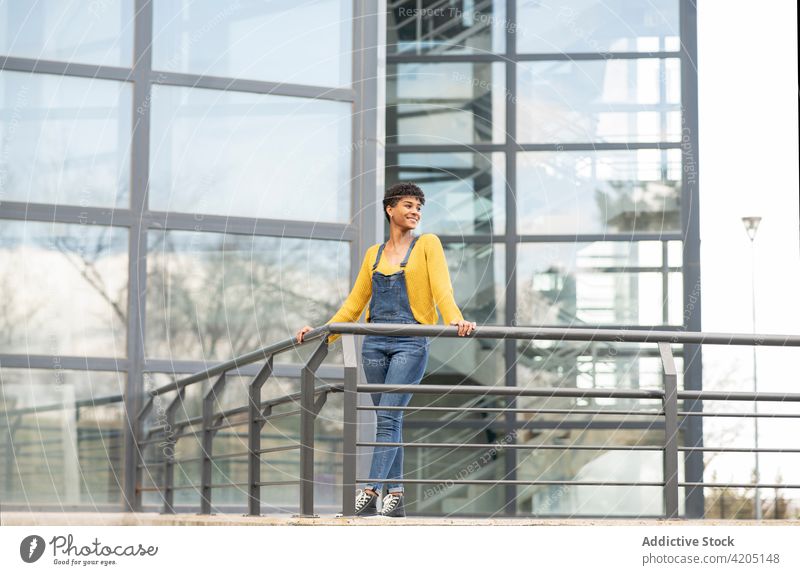 Lächelnde schwarze Frau steht in der Nähe eines modernen Gebäudes in der Stadt Großstadt urban trendy Outfit Stil Glaswand heiter cool ethnisch Afroamerikaner