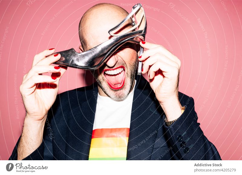 Ausdrucksstarker Schwuler mit Make-up, der einen hochhackigen Schuh im Studio vorführt Mann schwul Augen abdecken lgbt Homosexualität exzentrisch Konzept
