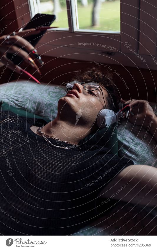 Schwuler mit Kopfhörern, der auf dem Bett im Bungalow mit seinem Smartphone chattet queer plaudernd Multimedia freie Zeit zuhören Musik benutzend Apparatur Mann