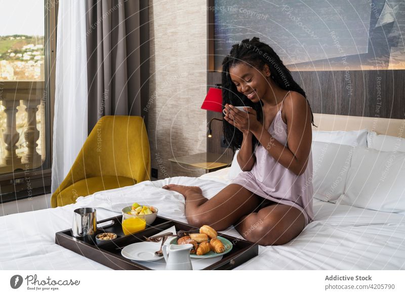 Glückliche junge schwarze Dame beim Frühstück in einem gemütlichen Hotelzimmer Frau trinken Kaffee Bett Inhalt Lächeln Komfort Tourist sich[Akk] entspannen