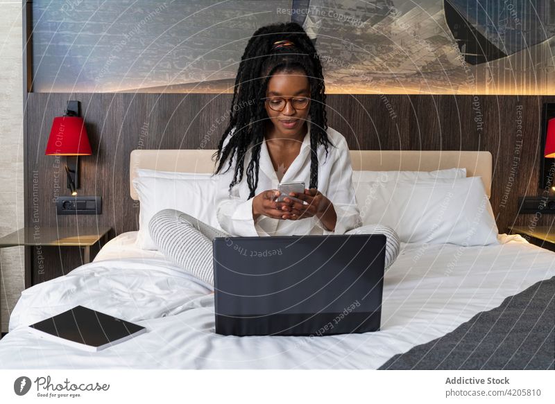 Selbstständige junge schwarze Frau mit Laptop und Smartphone auf dem Bett, während sie aus der Ferne arbeitet benutzend freiberuflich Arbeit beschäftigt