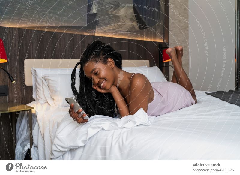 Fröhliche junge schwarze Dame mit Smartphone auf gemütlichem Bett Frau benutzend Glück Lächeln sich[Akk] entspannen Verlockung Nachricht soziale Netzwerke