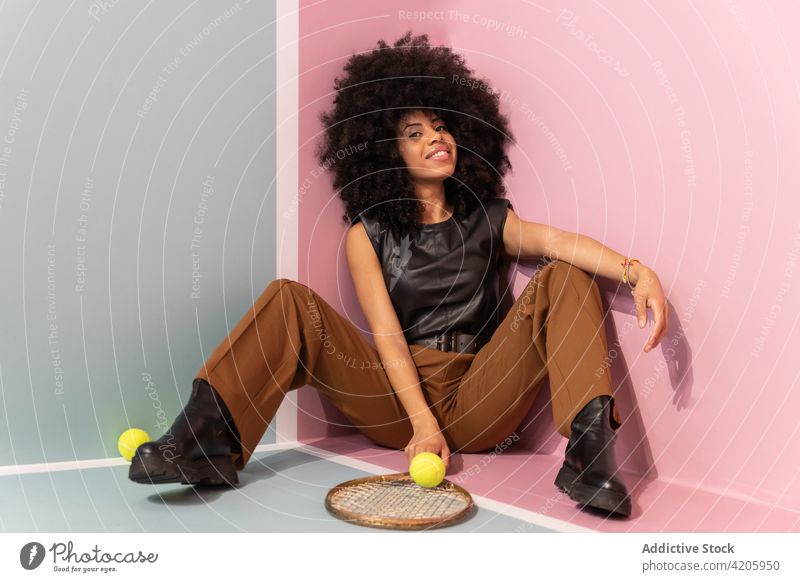 Schwarze Frau mit Tennisschläger sitzt im Studio Spieler Gericht Körperhaltung Remmidemmi Mode Atelier trendy Stil Afro-Look sitzen selbstbewusst Persönlichkeit