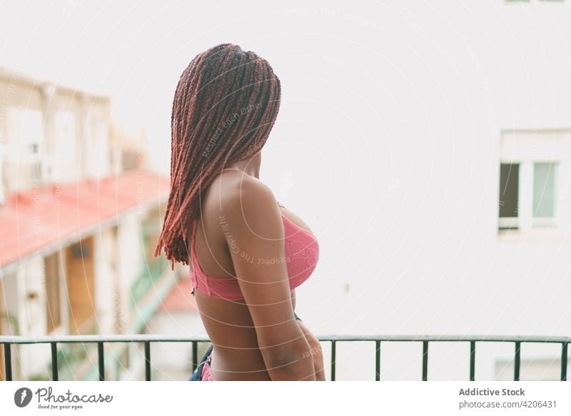 Schwarze Frau mit Zöpfen steht auf einem Balkon BH Geflecht Frisur beobachten Straße Dessous Unterwäsche Terrasse ethnisch schwarz Afroamerikaner rosa Stil