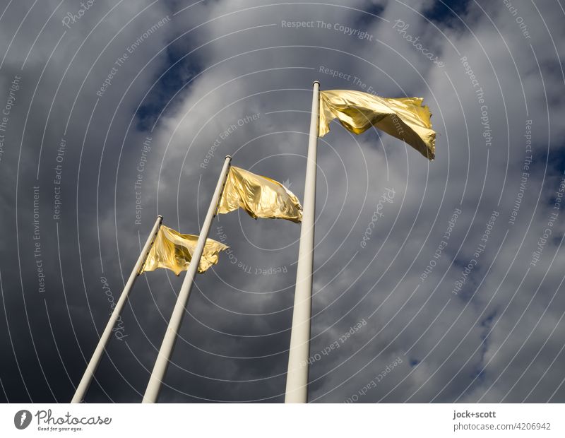 goldene Beflaggung flattert im Wind Fahnenmast Flagge Himmel Wolken wehen flattern Sonnenlicht Straßenkunst drei dunkler Hintergrund Froschperspektive