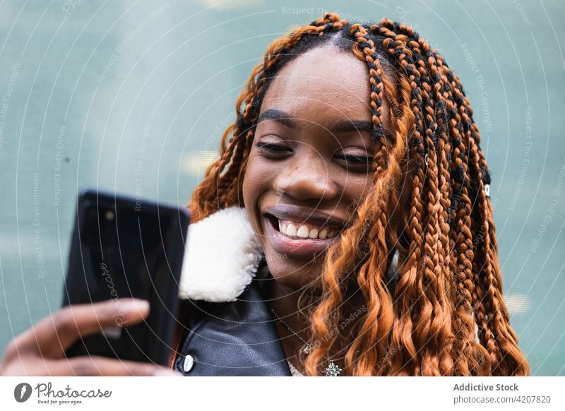 Stilvolle glückliche schwarze Frau benutzt Smartphone auf der Straße Browsen Glück Großstadt heiter trendy Outfit Geflecht Frisur soziale Netzwerke ethnisch