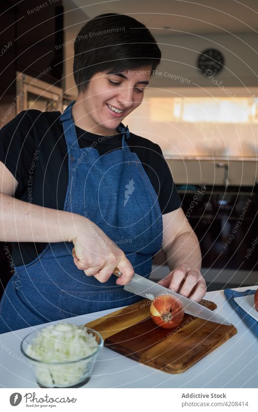 Glückliche Hausfrau beim Zwiebelschneiden in der Küche Frau geschnitten Koch heimwärts Lächeln organisch roh Schürze heiter kulinarisch frisch Schneidebrett