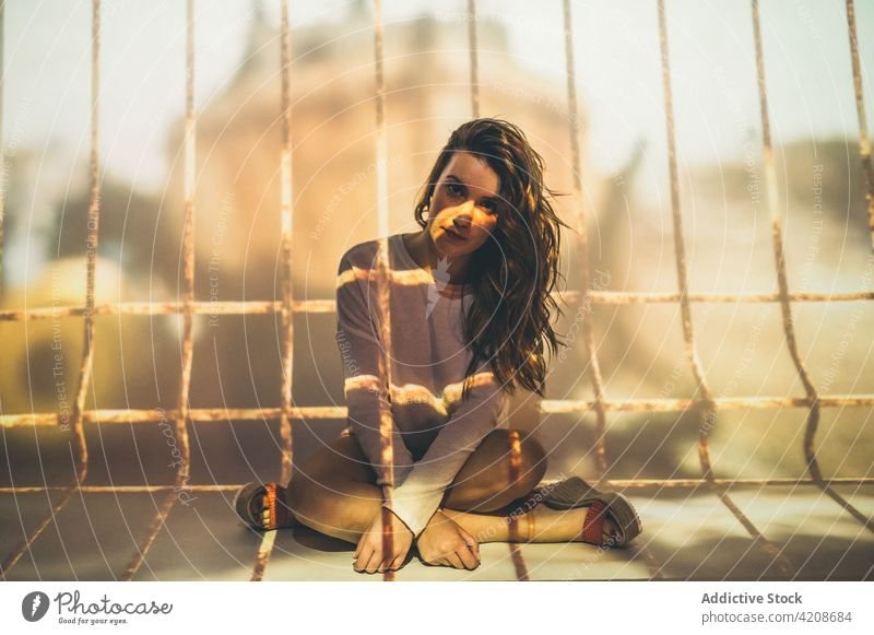 Junge Frau unter Projektion des Käfigs Konzept Gefangene Sommer Gitter Bar Outfit Beine gekreuzt Falle einsam allein Einsamkeit lange Haare lässig Gehege
