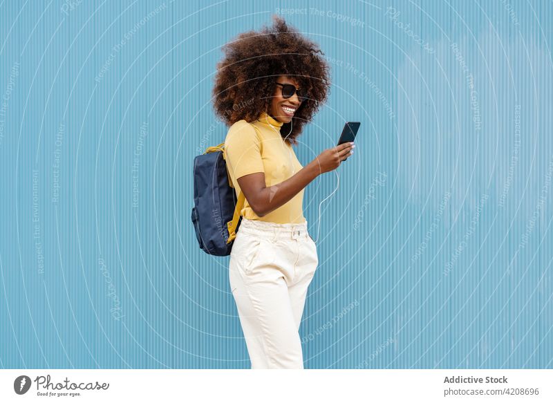 Fröhliche schwarze Frau, die in der Nähe einer blauen Wand auf ihrem Smartphone surft Browsen Selfie Moment Inhalt Kopfhörer Porträt benutzend Apparatur