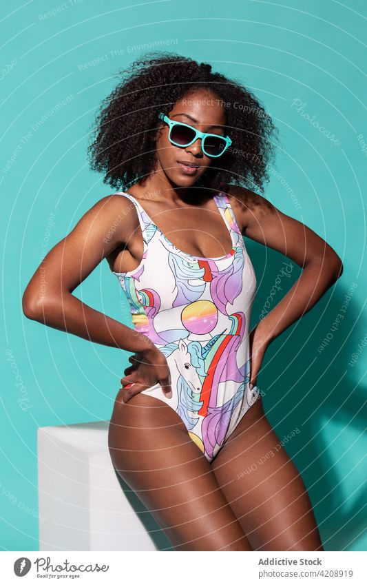 Wunderschöne schwarze Frau im Badeanzug sitzt im hellen Studio Badebekleidung Stil Hand auf der Taille Körperhaltung heiter Outfit Sonnenbrille cool