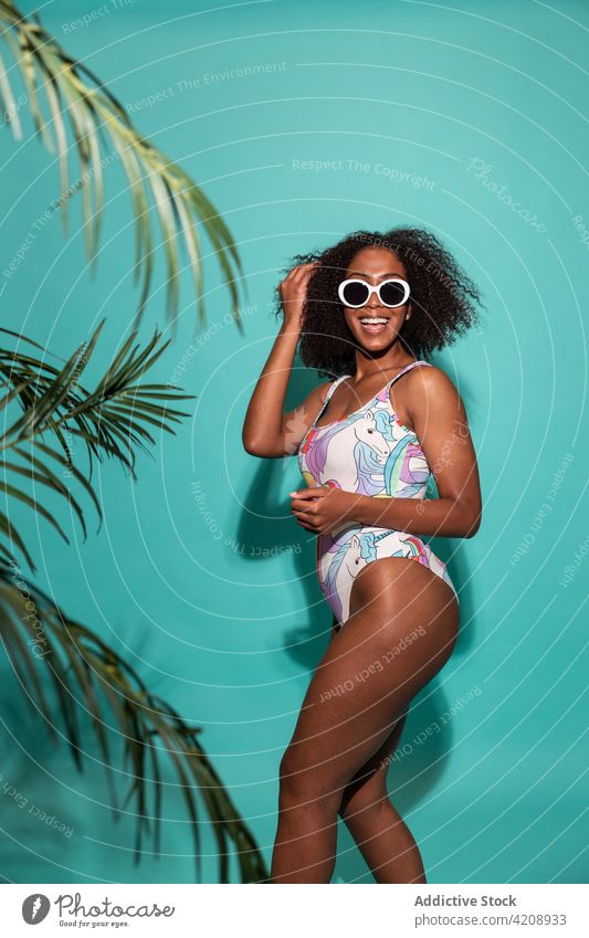 Fröhliche schwarze Frau im Badeanzug auf blauem Hintergrund Badebekleidung Stil Glück Haare berühren heiter Sonnenbrille cool charismatisch Freude Outfit