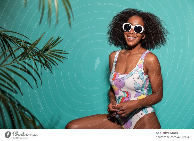 Fröhliche schwarze Frau im Badeanzug sitzt auf blauem Hintergrund Badebekleidung Stil Glück heiter Sonnenbrille cool charismatisch Freude Outfit sorgenfrei