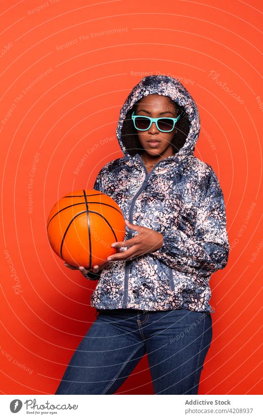 Selbstbewusste schwarze Frau steht mit Basketball im Studio selbstbewusst Ball Stil cool Outfit sportlich Streetstyle Sonnenbrille Vorschein Accessoire ernst