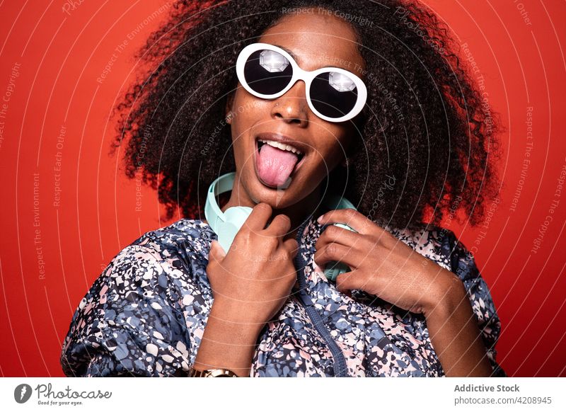 Stilvolle schwarze Frau mit Kopfhörern auf rotem Hintergrund cool ausspannen Musik Arme hochgezogen Outfit Grimasse Gesicht machen Glück unterhalten Gesang
