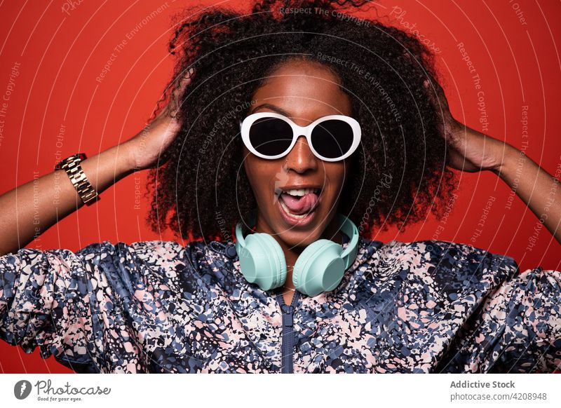 Stilvolle schwarze Frau mit Kopfhörern berühren Haare auf rotem Hintergrund cool ausspannen Musik Arme hochgezogen Haare berühren Outfit Grimasse Gesicht machen