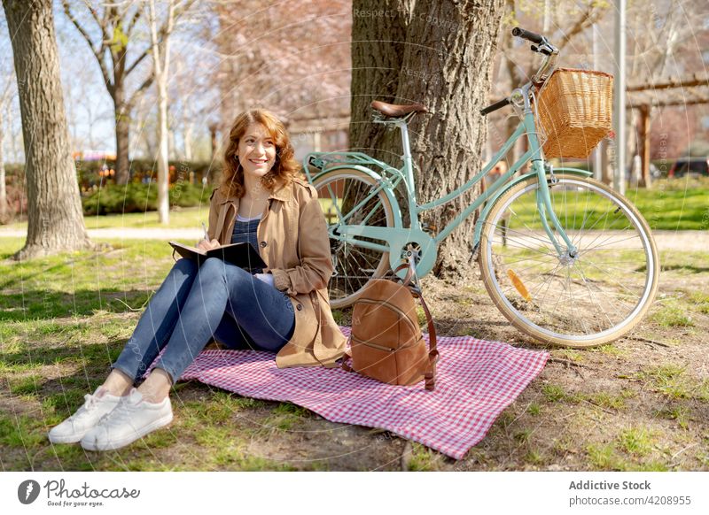 Glückliche junge Frau, die auf einer Wiese neben einem Fahrrad in einen Planer schreibt schreiben Notebook Park zur Kenntnis nehmen kariert Gewebe nachdenklich