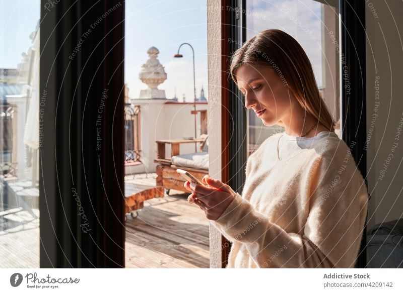 Nachdenkliche junge Frau, die ihr Smartphone in der Nähe der Terrasse ihres Hauses benutzt Nachricht Fenster heimwärts Kommunizieren Talkrunde Internet online