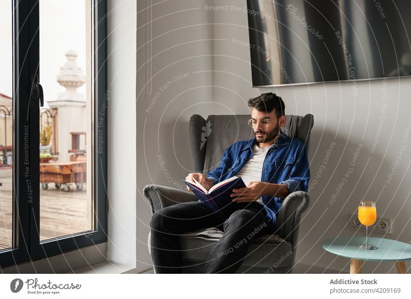 Ernster junger ethnischer Mann sitzt im Sessel und liest einen Roman lesen Buch Armsessel heimwärts sich[Akk] entspannen Wissen Literatur Hobby Konzentration