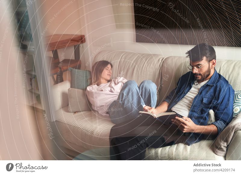 Ernster hispanischer Mann, der auf dem Sofa in der Nähe seiner schlafenden Freundin ein Buch liest Paar ruhen lesen Literatur Partnerschaft Zusammensein